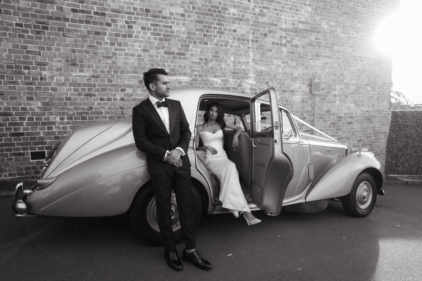 A bride and groom beside a vintage wedding car in black and white